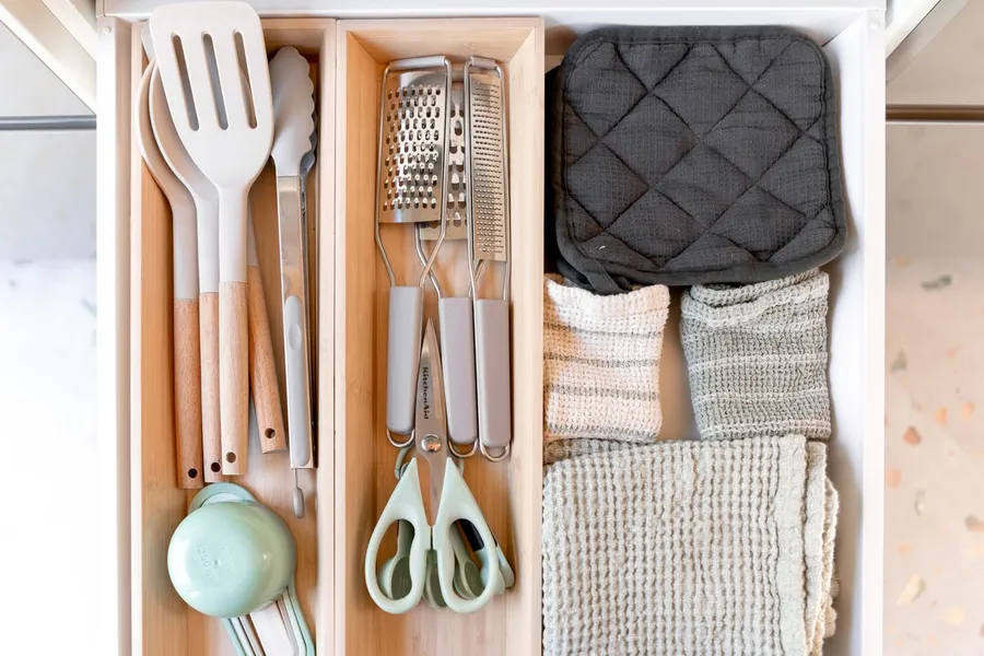 Open kitchen drawer with wooden dividers, cooking utensils, graters, scissors, and folded kitchen towels
